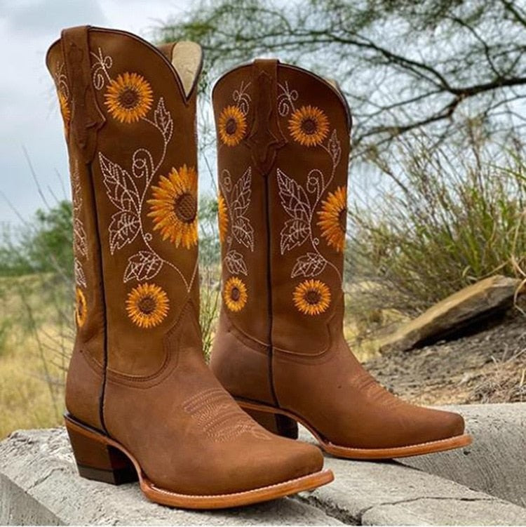 Dunkelbraune Cowboystiefel mit Sonnenblumen-Stickerei vor Holzzaun
